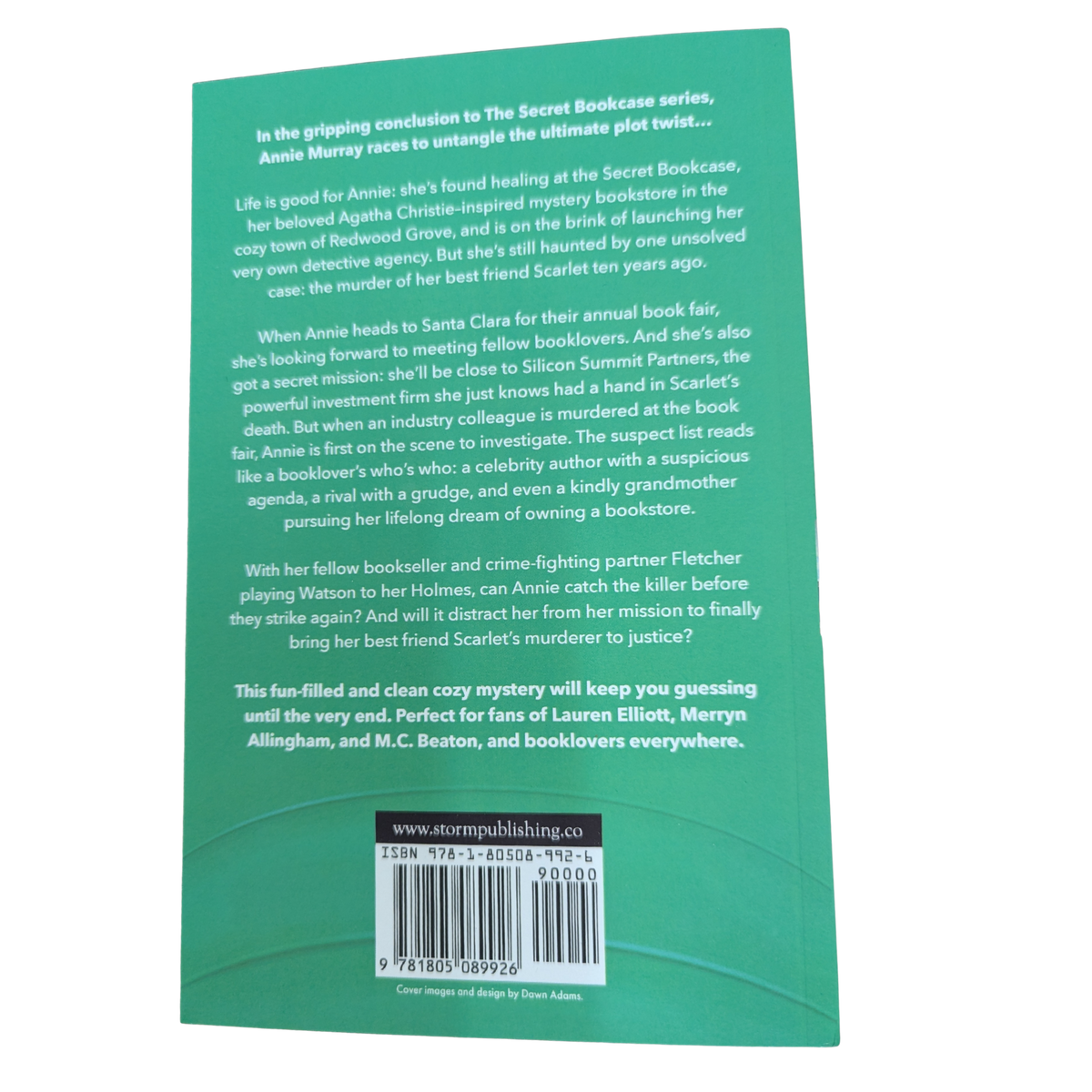 A Body at the Book Fair by Ellie Alexander cozy mystery paperback cover featuring a colorful outdoor book fair with book stalls, stacked books, trees, and a fountain.
