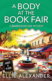 A Body at the Book Fair by Ellie Alexander cozy mystery paperback cover featuring a colorful outdoor book fair with book stalls, stacked books, trees, and a fountain.