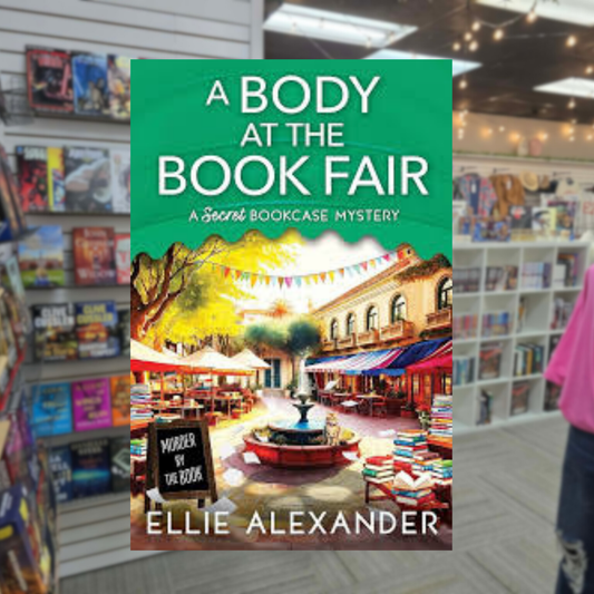A Body at the Book Fair by Ellie Alexander cozy mystery paperback cover featuring a colorful outdoor book fair with book stalls, stacked books, trees, and a fountain.