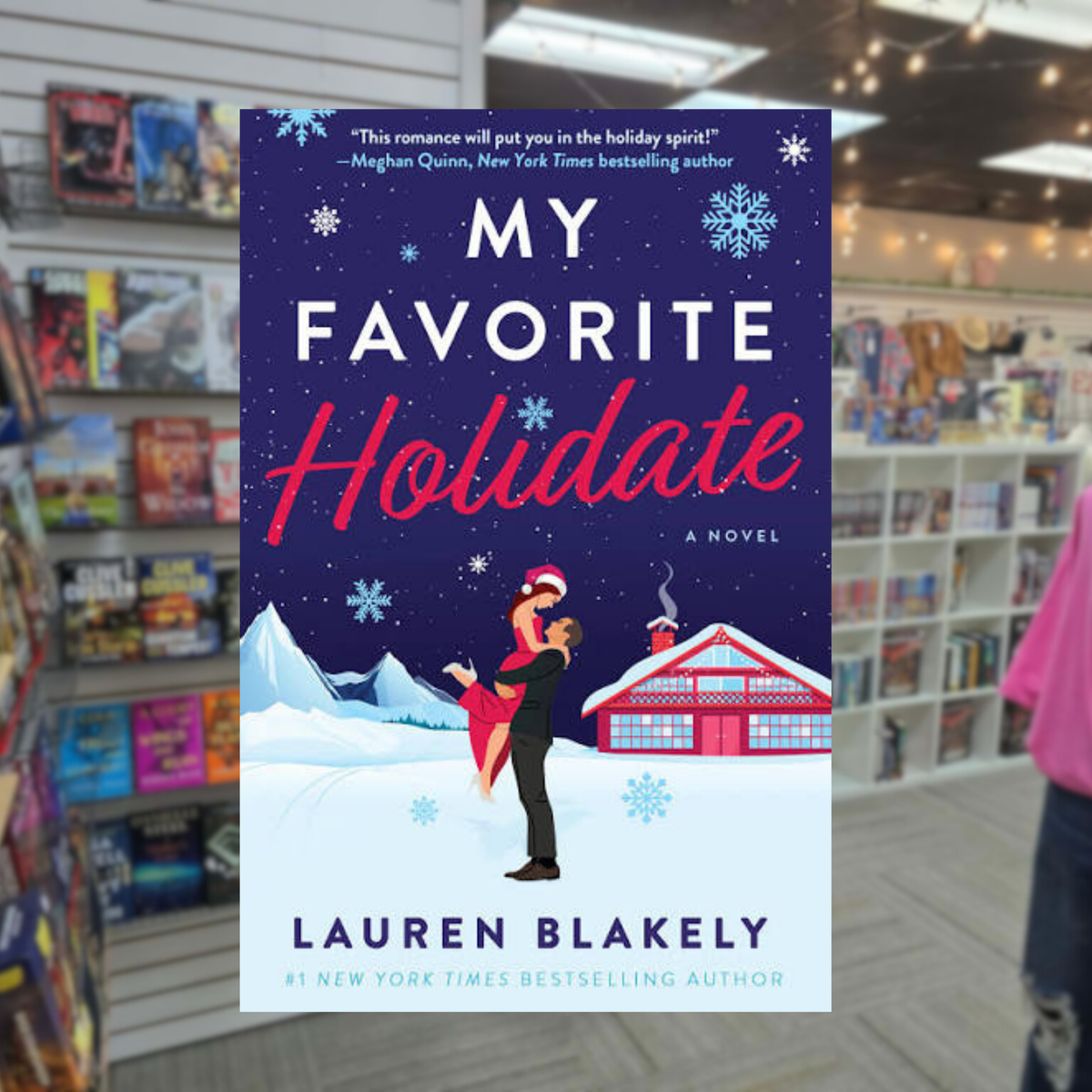 My Favorite Holidate by Lauren Blakely Christmas romance paperback cover featuring a couple embracing in a snowy mountain setting beside a festive red cabin, with snowflakes against a dark blue sky.