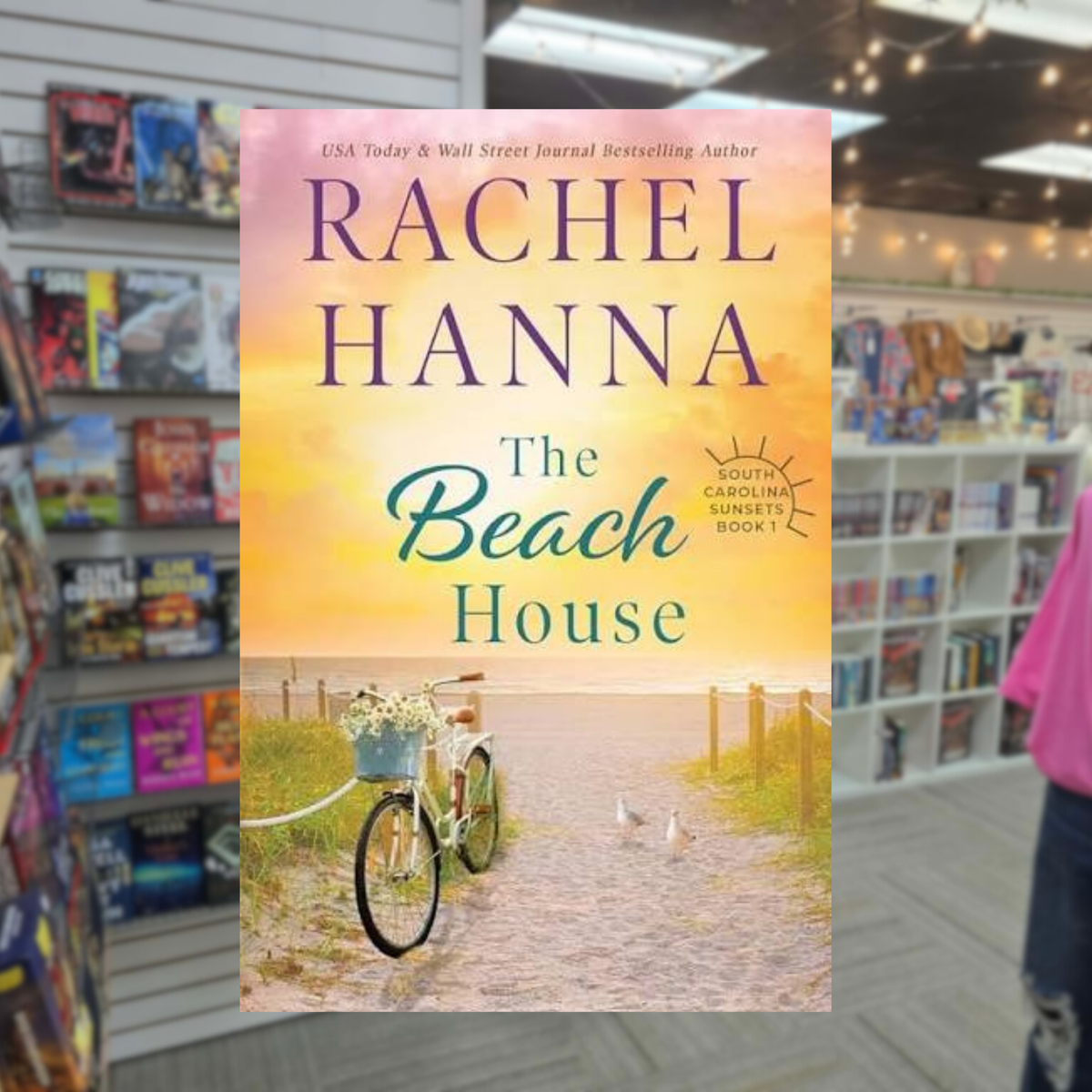 The Beach House by Rachel Hanna paperback cover featuring a bicycle with a basket of flowers on a sandy path leading to the beach at sunset.