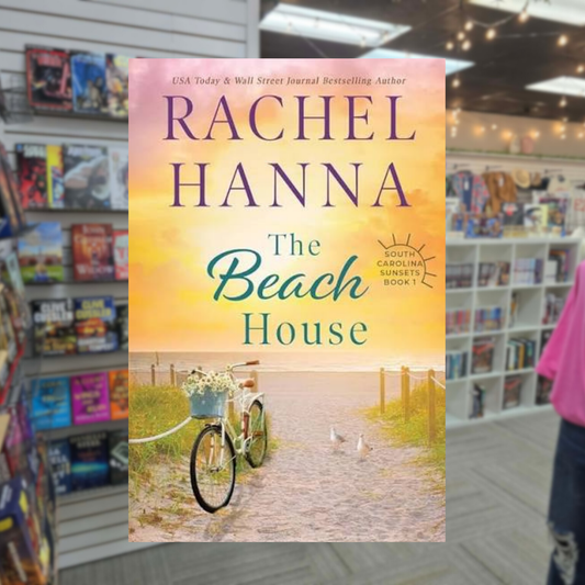 The Beach House by Rachel Hanna paperback cover featuring a bicycle with a basket of flowers on a sandy path leading to the beach at sunset.