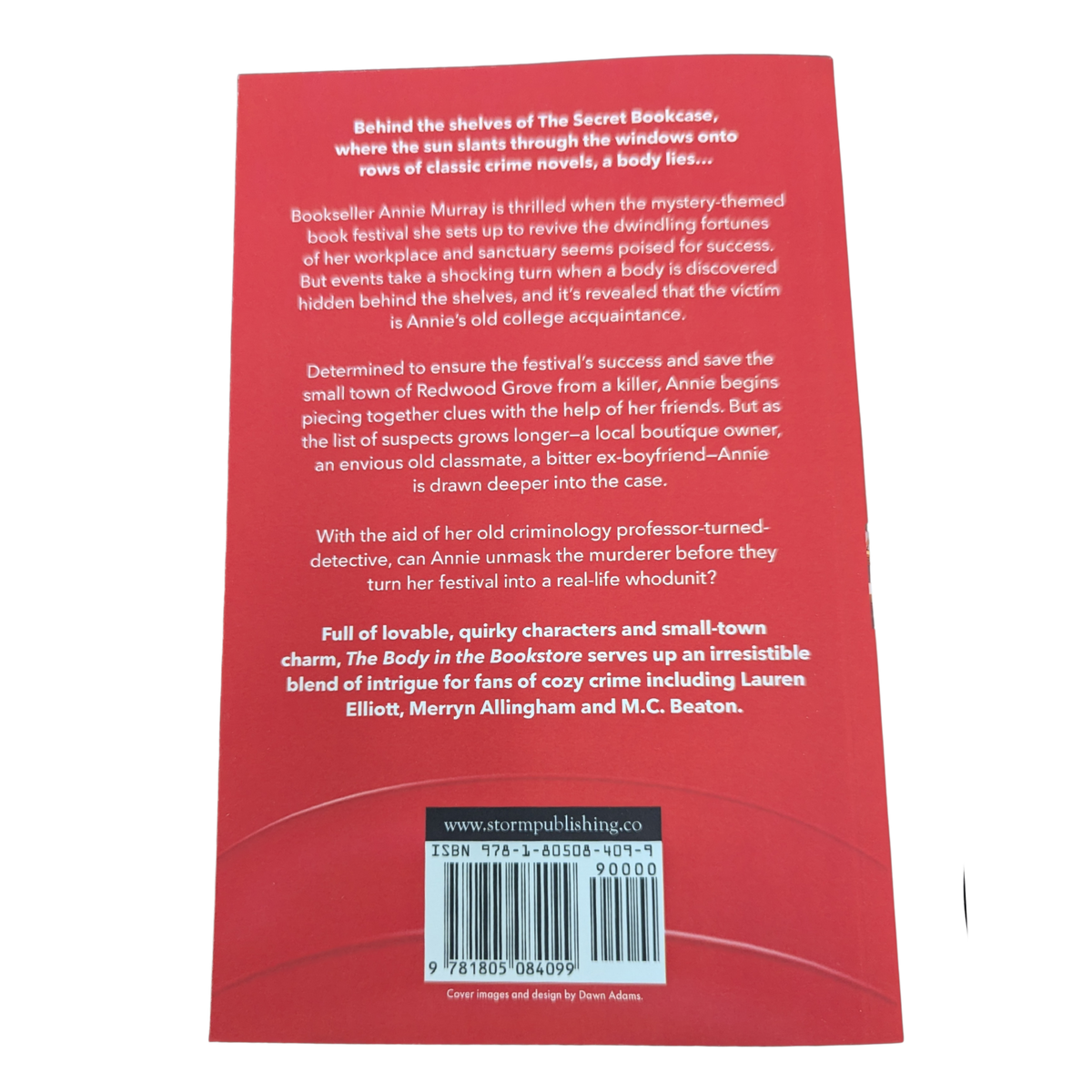 The Body in the Bookstore by Ellie Alexander cozy mystery paperback cover featuring a charming bookstore interior with bookshelves, a reading table, large windows, and red curtains.