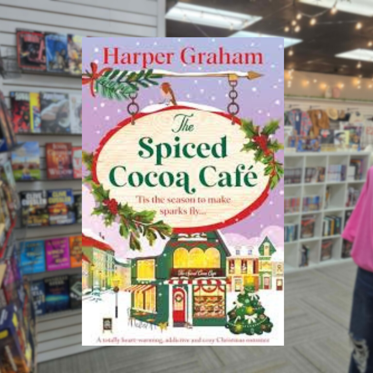The Spiced Cocoa Café by Harper Graham Christmas romance book cover featuring a charming festive café, snow-covered buildings, Christmas greenery, colorful holiday decorations, and a snowy winter village.